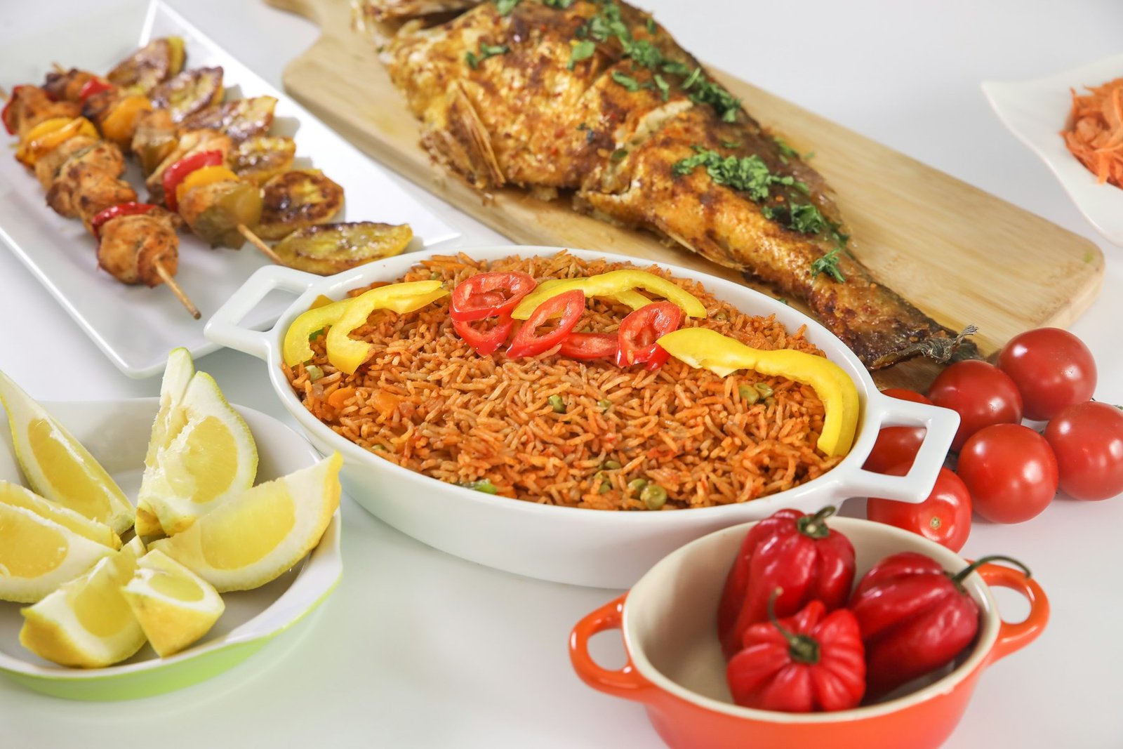Jollof rice in a white dish with grilled fish, skewers and fresh vegetables
